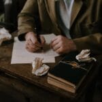 Osoba pisze przy drewnianym biurku, obok zmięte kartki i otwarta książka